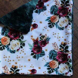 May include: A white blanket with a floral pattern featuring burgundy, orange, and cream-colored roses, green leaves, and small yellow accents. The blanket has a dark green, plush border.