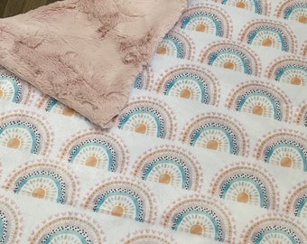 May include: A soft, plush blanket with a white background and a repeating pattern of colorful rainbows with a sun in each one. The blanket has a pink faux fur backing.