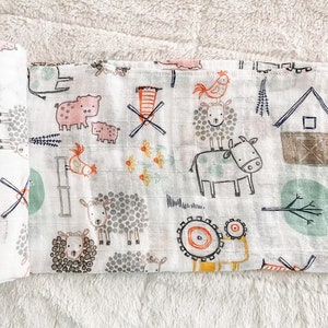 Op de afbeelding: Een witte katoenen muslin spuugdoek met een boerderijdierenprint. De print bevat varkens, kippen, schapen, koeien en een windmolen. De stof is zacht en absorberend.