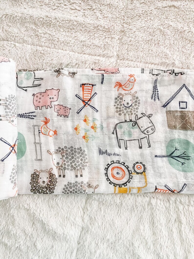 Op de afbeelding: Een witte katoenen muslin spuugdoek met een boerderijdierenprint. De print bevat varkens, kippen, schapen, koeien en een windmolen. De stof is zacht en absorberend.