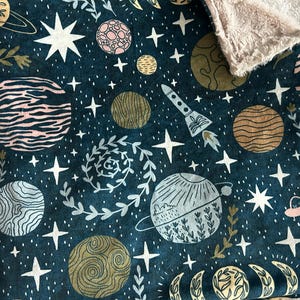 May include: A dark teal baby blanket with a space-themed pattern. The design includes planets, stars, a rocket ship, and a soft, tan-colored plush backing. The blanket is rectangular.
