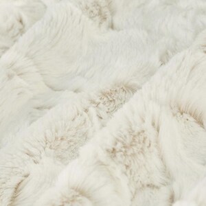 May include: Close-up of a plush, textured blanket in cream and light brown tones. The fabric has a soft, fur-like appearance with a subtle, quilted pattern. The blanket's surface is a mix of light and darker shades, creating depth.