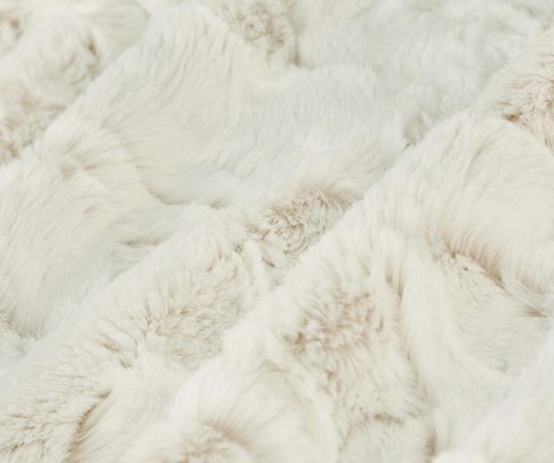 May include: Close-up of a plush, textured blanket in cream and light brown tones. The fabric has a soft, fur-like appearance with a subtle, quilted pattern. The blanket's surface is a mix of light and darker shades, creating depth.