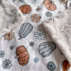 May include: A soft, cream-colored throw blanket with a repeating pattern of orange, brown, and gray pumpkins and white daisies. The blanket has a fluffy, faux fur backing.