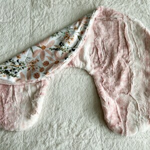 以下が含まれることがあります： 花柄のピンクと白のベビー用おくるみ。おくるみは柔らかく、ふわふわした生地で作られており、赤ちゃんを暖かく快適に保つように設計されています。