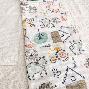 Op de afbeelding: Een witte muslin-wikkeldeken met een boerderijdierenprint. De print toont koeien, schapen, kippen, een tractor en een windmolen. De deken is zacht en ademend, waardoor het perfect is om een ​​baby in te wikkelen.