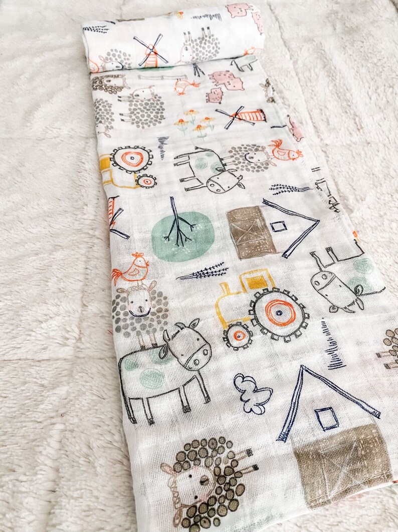 Op de afbeelding: Een witte muslin-wikkeldeken met een boerderijdierenprint. De print toont koeien, schapen, kippen, een tractor en een windmolen. De deken is zacht en ademend, waardoor het perfect is om een ​​baby in te wikkelen.
