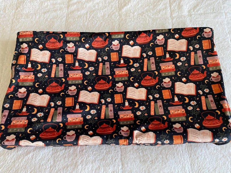May include: A dark blue fabric with a repeating pattern of teapots, books, teacups, candles, moons, and flowers in shades of red, orange, and white. The fabric is likely used for crafting or home decor.