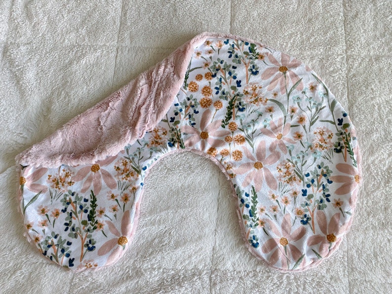 以下が含まれることがあります： ピンクと白の花柄の授乳枕で、柔らかくふわふわのピンクの面があります。枕は三日月型で、授乳中または哺乳瓶で赤ちゃんを支えるように設計されています。