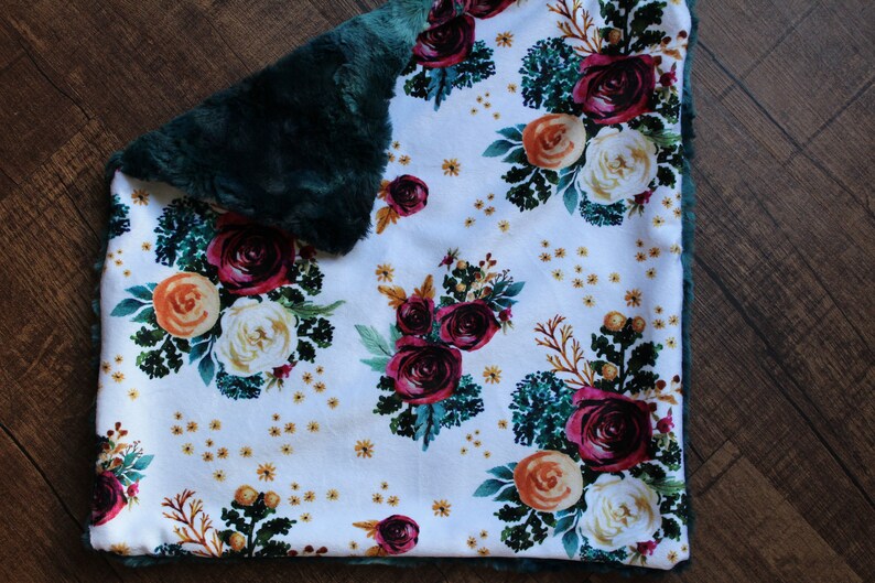 May include: A white blanket with a floral pattern featuring burgundy, orange, and cream-colored roses, green leaves, and small yellow accents. The blanket has a dark green, plush border.