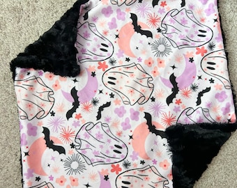 May include: A white and pink fleece blanket with a black fleece backing. The blanket features a repeating pattern of ghosts, bats, moons, and flowers.