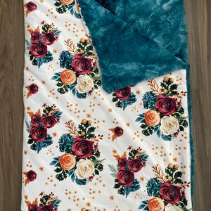 May include: A white blanket with a floral pattern featuring burgundy, peach, and cream roses, accented with teal foliage and small yellow flowers. The blanket has a teal, plush, furry backing.