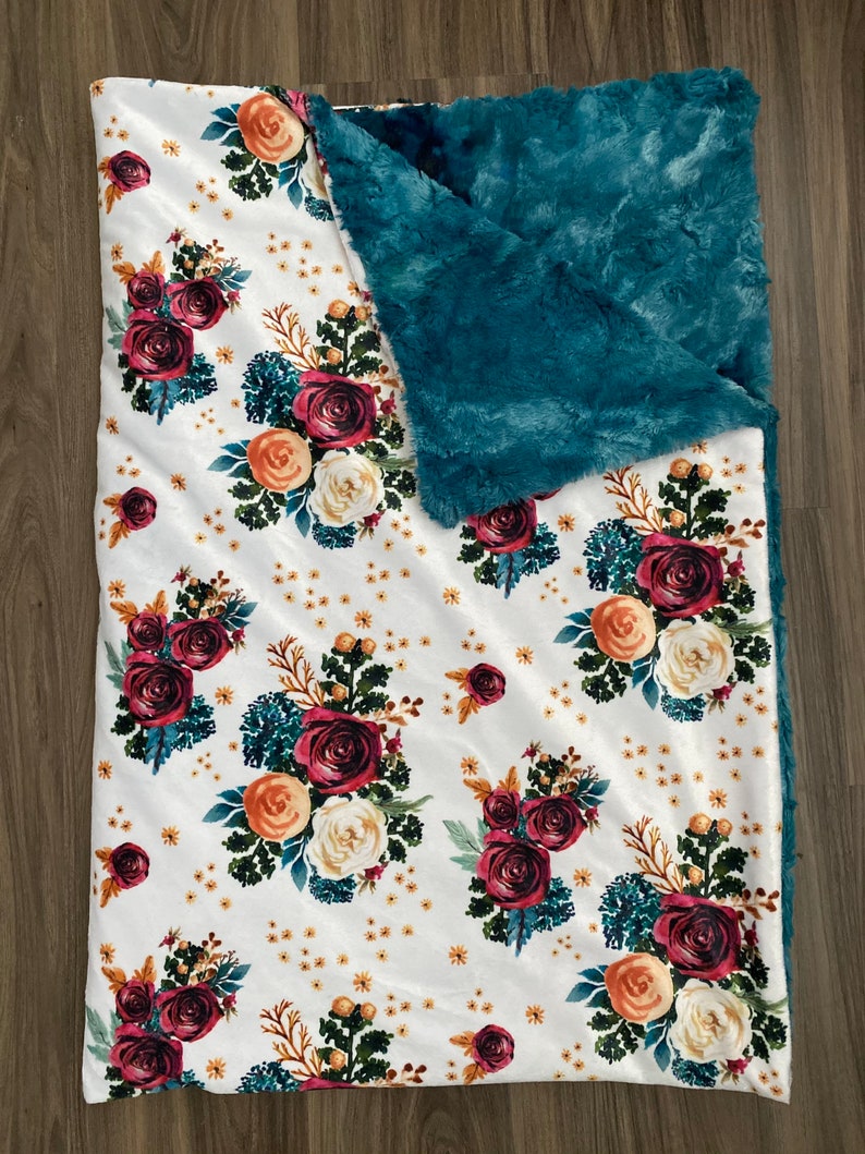 May include: A white blanket with a floral pattern featuring burgundy, peach, and cream roses, accented with teal foliage and small yellow flowers. The blanket has a teal, plush, furry backing.