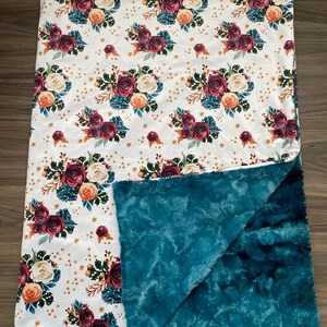 May include: A soft, floral-patterned blanket with a white top featuring burgundy, cream, and peach roses, teal foliage, and gold accents. The underside is a plush teal fabric, creating a cozy and decorative item.