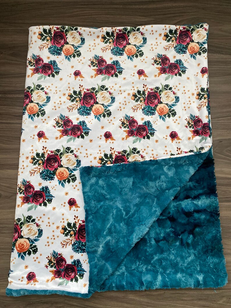 May include: A soft, floral-patterned blanket with a white top featuring burgundy, cream, and peach roses, teal foliage, and gold accents. The underside is a plush teal fabric, creating a cozy and decorative item.