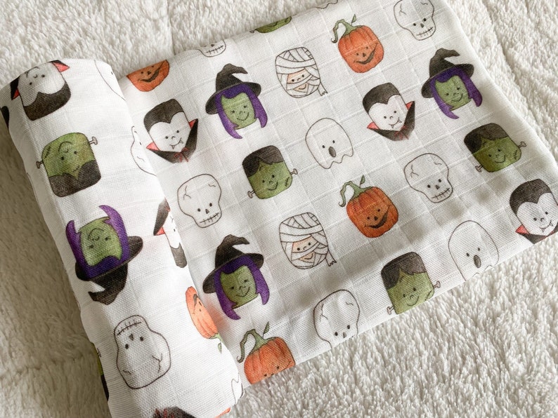 May include: White Halloween-themed baby swaddle blanket featuring a repeating pattern of cartoon pumpkins, vampires, witches, mummies, ghosts, Frankenstein's monster, and skulls. The blanket is rolled up and displayed on a white, textured surface.