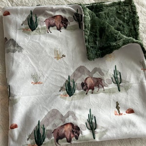 May include: A white and green baby blanket with a buffalo and cactus print. The blanket has a soft, plush green backing.