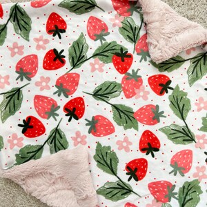 May include: A white baby blanket with a pink minky backing. The blanket features a repeating pattern of red strawberries, green leaves, and pink flowers.