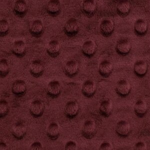 May include: Close-up of a burgundy minky fabric with a raised dot pattern. The plush material has a soft, velvety texture and a deep, rich color. The dots are evenly spaced across the surface, creating a tactile and visually appealing design.