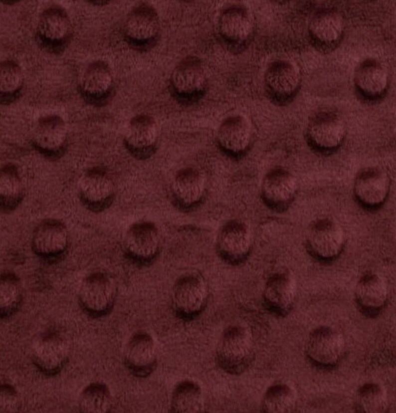 May include: Close-up of a burgundy minky fabric with a raised dot pattern. The plush material has a soft, velvety texture and a deep, rich color. The dots are evenly spaced across the surface, creating a tactile and visually appealing design.