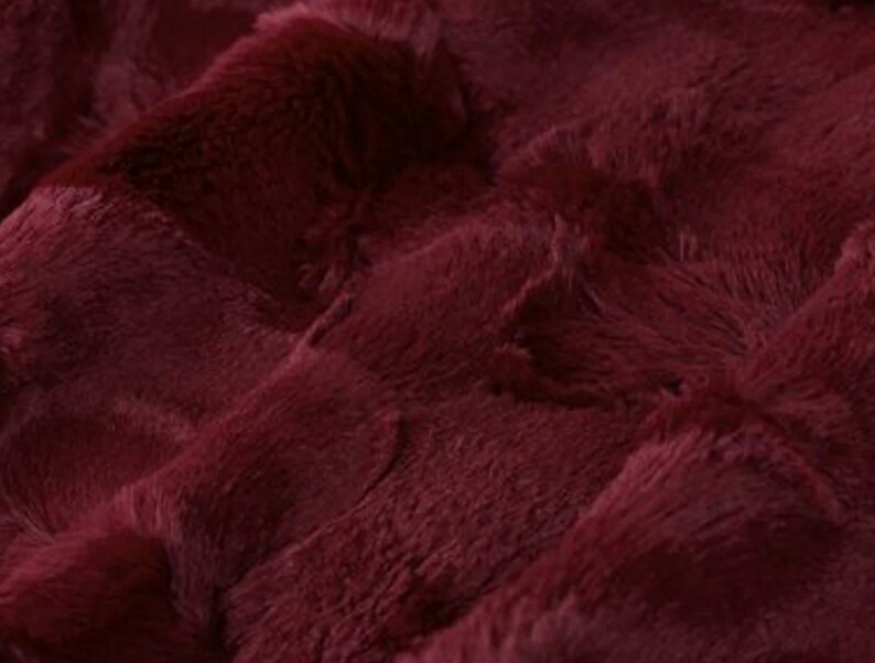 May include: Close-up of a plush, burgundy fabric with a textured, rippled surface. The material has a soft, furry appearance, with visible fibers and a deep, rich color. The fabric's folds create shadows and highlights.