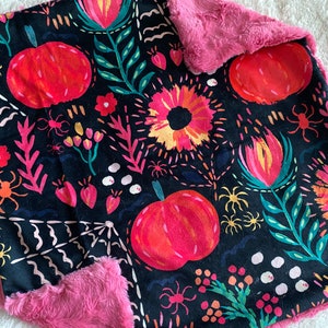 May include: A black and pink fleece blanket with a Halloween-themed print featuring pumpkins, flowers, spiders, and leaves. The print is in shades of pink, orange, yellow, green, and white.