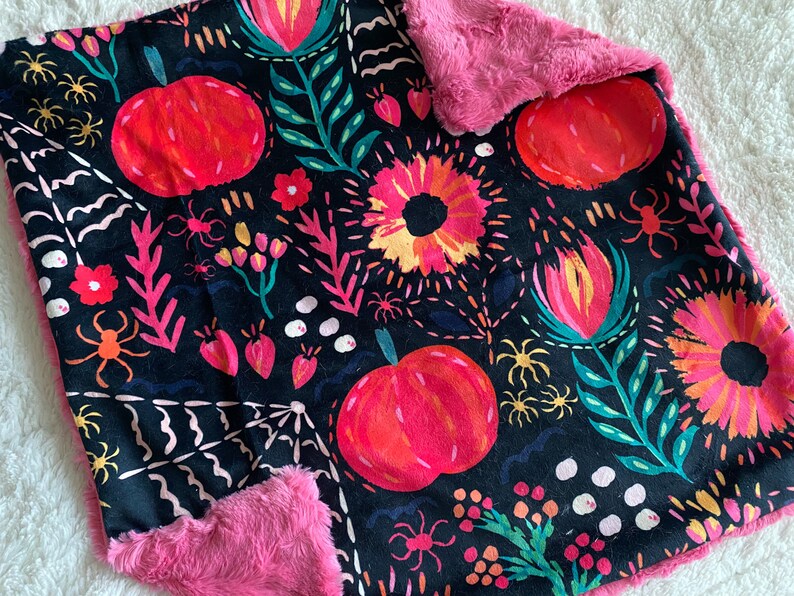 May include: A black and pink fleece blanket with a Halloween-themed print featuring pumpkins, flowers, spiders, and leaves. The print is in shades of pink, orange, yellow, green, and white.
