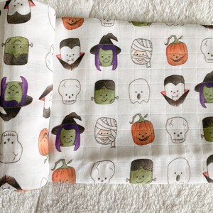 May include: White baby blanket with a repeating pattern of Halloween characters, including ghosts, vampires, witches, pumpkins, skulls, and Frankenstein's monster. The characters are in shades of orange, green, purple, and black.