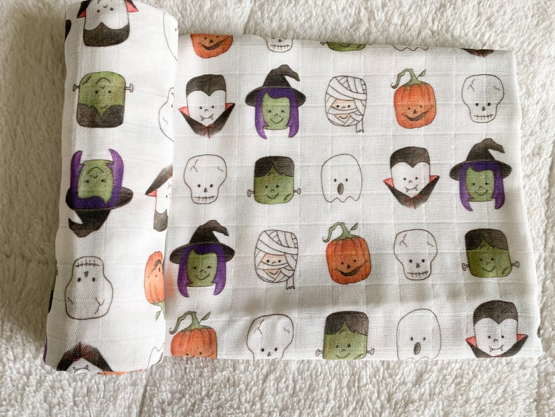May include: White baby blanket with a repeating pattern of Halloween characters, including ghosts, vampires, witches, pumpkins, skulls, and Frankenstein's monster. The characters are in shades of orange, green, purple, and black.