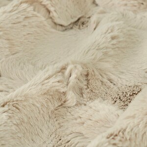 May include: Close-up of a plush, faux fur fabric in a neutral beige color. The fabric has a dense, textured surface with long, soft fibers, creating a luxurious and cozy appearance. The folds and shadows add depth to the material.