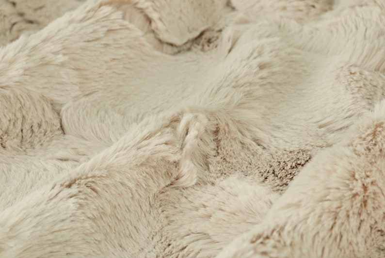 May include: Close-up of a plush, faux fur fabric in a neutral beige color. The fabric has a dense, textured surface with long, soft fibers, creating a luxurious and cozy appearance. The folds and shadows add depth to the material.