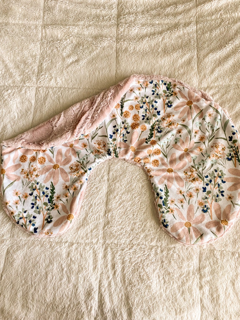 以下が含まれることがあります： ピンク、白、オレンジの花が咲いた花柄のU字型の妊娠用枕。枕は柔らかく、ふわふわした生地で覆われています。