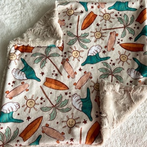 May include: A cream-coloured baby blanket featuring a repeating pattern of surfboards, palm trees, smiling suns, and ocean waves in orange, teal, and beige. The blanket has a soft, plush, beige border.