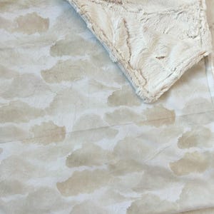 May include: A soft, plush blanket with a cloud-like pattern in shades of beige and white. The blanket features a textured, fluffy border and a smooth, printed interior. The design creates a cozy and inviting aesthetic, perfect for home decor.