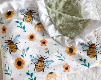 May include: A white baby blanket with a green minky backing and a white satin trim. The blanket features a repeating pattern of bees and sunflowers in watercolor style.