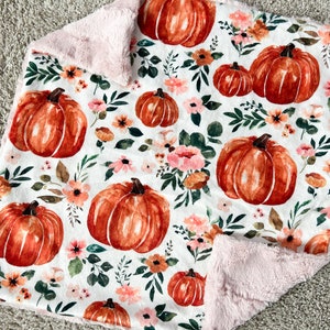 May include: A soft, white baby blanket with a pink fleece backing. The blanket features a watercolor pattern of orange pumpkins and pink flowers.