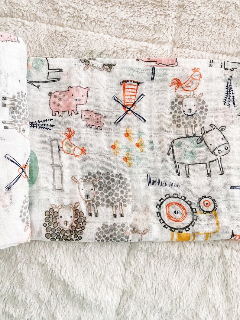 Op de afbeelding: Een witte katoenen spuugdoek met een boerderijdierenprint. De print bevat varkens, schapen, koeien, kippen en windmolens in zwart, bruin, roze, oranje en groen.