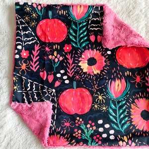 Op de afbeelding: Een roze en zwarte babydeken met een bloemen- en pompoenprint. De deken is gemaakt van zacht, pluche stof.