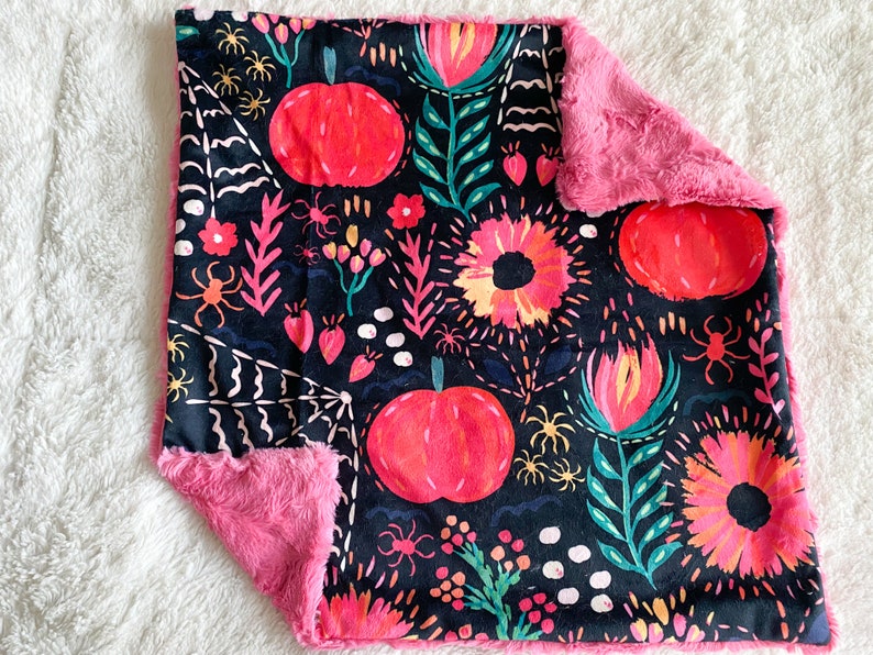Op de afbeelding: Een roze en zwarte babydeken met een bloemen- en pompoenprint. De deken is gemaakt van zacht, pluche stof.