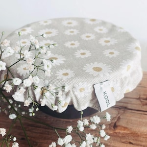 Op de afbeelding: Een grijze en witte stoffen komdeksel met een print van madeliefjes en een wit label met de tekst "KOOL" erop. Het deksel ligt op een houten oppervlak.