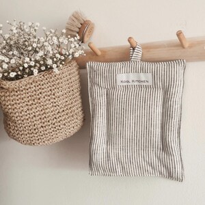 Peut inclure: Un manique rectangulaire en tissu à rayures verticales grises et blanches, doté d'une petite étiquette blanche avec le texte KOOL KITCHEN. Le manique possède une boucle en tissu assortie pour le suspendre à un crochet mural en bois.