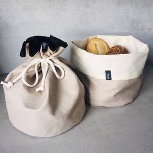 Puede incluir: Dos bolsas de lona beige para guardar pan. Una bolsa es una bolsa de cordón con un ribete negro y el texto "Griffe" en el ribete. La otra bolsa es una cesta rectangular con un forro blanco y el texto "Kook" en el fondo. Ambas bolsas están llenas de bollos.