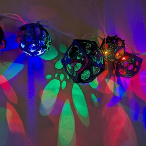 May include: A string of decorative lights with black, geometric, cage-like bulbs. The lights cast colorful shadows on the wall, including shades of green, blue, and red.