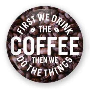 May include: A round button with a brown coffee bean background. The text on the button reads "First we drink the coffee then we do the things".
