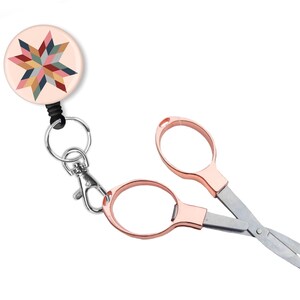 May include: A pink and gold retractable badge holder with a colorful starburst design and a pair of small rose gold scissors attached to a silver key ring.