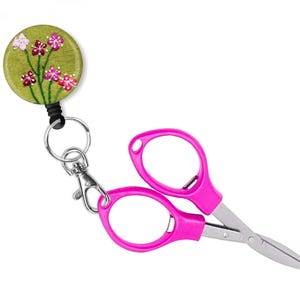 May include: A pink pair of small scissors with a retractable cord and a green button with embroidered flowers. The button is attached to a silver key ring.