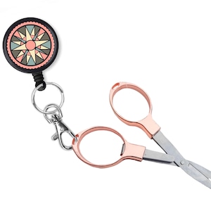 May include: A rose gold colored pair of small scissors with a retractable keychain. The keychain has a black retractable cord and a round black and white design with a pink starburst in the center.