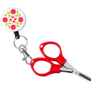 May include: A pair of red-handled scissors with a floral design retractable badge. The scissors have a silver blade and are attached to a badge with a white background and a pink and green floral pattern.