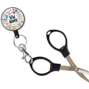 May include: A retractable badge reel with a "SEW HAPPY" design and a pair of black-handled curved scissors. The reel features sewing-themed illustrations. The scissors have silver blades and are open.