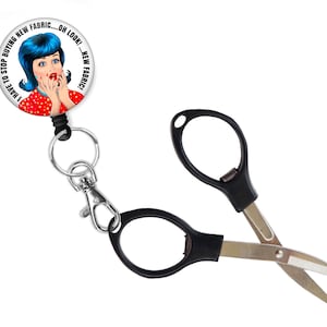 May include: A retractable badge reel with a white button featuring a cartoon woman with blue hair and the text "I HAVE TO STOP BUYING NEW FABRIC... OH LOOK! ... NEW FABRIC". The reel is attached to a pair of black and silver small scissors.